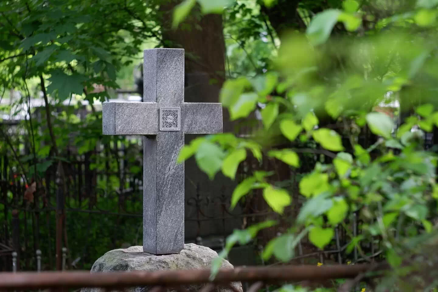 Кам'яний хрест з меморіальною табличкою з QR-кодом на тлі зеленого цвинтарного пейзажу